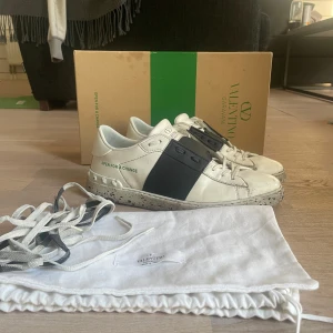 Valentino Garavani Open  - Snygga Valentino Garavani Open sneakers i vitt skinn med bred svart läderdetalj över sidan. Sulan är vit med svarta stänk och har en platt form. Skorna har snörning och rund tå, samt logga på plösen. Medföljer extra skosnören och dustbag. Nypris 7200kr. Fint skick ända dåliga är på bild 4. Pris kan diskuteras! Hör av för minsta fundering! 