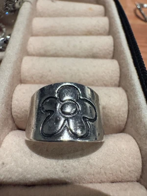 Bred silverfärgad ring med blomma - Cool bred ring i silverfärgad metall med ett ingraverat blommotiv framtill. Ringen är öppen baktill så storleken kan justeras. Perfekt statement-piece för dig som gillar retrovibbar och unika accessoarer.