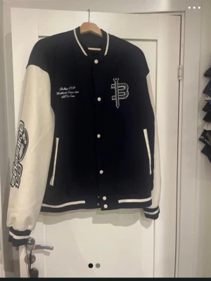Varsityjacka från ASOS, storlek XL - Svart och vit varsityjacka från Asos med broderade detaljer och tryck på både fram- och baksida. Jackan har vita ärmar, ribbade muddar och knappar framtill. Snygg kontrast mellan färgerna och coolt motiv på ryggen. Storlek XL, använd nån enstaka gång