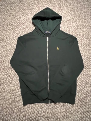 Mörkgrön Ralph Lauren Hoodie  - Säljer en mörkgrön hoodie från Ralph Lauren i storlek Small. Tröjan är i mycket bra skick med minimalt slitage och inga fläckar, hål eller noppor. Klassisk modell med liten broderad logga.                       Bredd: 45 Längd: 60