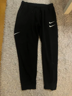 Svarta Nike mjukisbyxor med tryck - Svarta mjukisbyxor från Nike med vita Swoosh-loggor på benen och röd text på bakfickan. Byxorna har snörning i midjan och muddar vid bensluten. Tillverkade i mjukt bomullsmaterial, perfekt för chill eller träning. Byxorna har några små igensydda hål.
