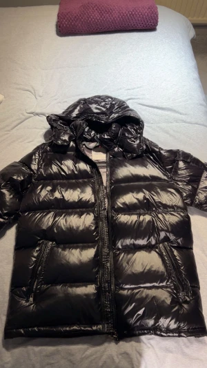 Svart Moncler pufferjacka med huva - Säljer en svart, glansig pufferjacka från Moncler med huva och dragkedja framtill. Jackan har två snedställda fickor med dragkedja och en ficka med knapp på ärmen där Moncler-loggan sitter. Insidan är quiltad och jackan har en loose passform.