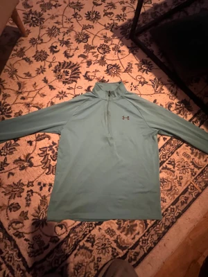Turkos långärmad träningströja Under Armour - Snygg turkos långärmad träningströja från Under Armour med halv dragkedja och hög krage. Tröjan har logga på bröstet och ryggen, loose fit och är tillverkad i mjukt syntetmaterial som andas. Perfekt för gymmet eller löprundan.