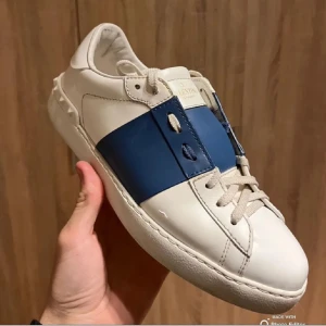 Valentino Garavani Open  - Snygga Valentino Garavani Open sneakers i vitt skinn med bred blå läderdetalj över mitten. Klassisk rund tå, platt sula och coola nitar baktill. Skorna har snörning och kommer med original dustbags. Perfekta för dig som vill sticka ut med stil.