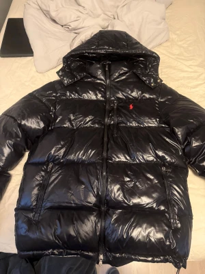 Svart pufferjacka från Polo Ralph Lauren - Säljer en svart pufferjacka från Polo Ralph Lauren med glansig finish, huva och flera dragkedjefickor. Jackan har det klassiska röda Polo-logot broderat på bröstet och RL-detaljer på dragkedjorna. Perfekt för kalla dagar och riktigt snygg streetstil. Jackan är aldrig använd men har inte lapp kvar då jag kastade taggen men original förpackning finns 