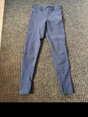 Blåa ridbyxor Alequi - Snygga blåa ridbyxor från Alequi med silikonprint för extra grepp och praktisk ficka på benet. Byxorna har en tight passform och är tillverkade i ett stretchigt syntetmaterial som ger bra rörelsefrihet. Perfekta för dig som vill ha stil och funktion i stallet.