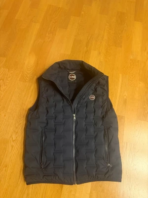 Mörkblå quiltad dunväst från Colmar - Mörkblå dunväst från Colmar med quiltad design och krage. Västen har två fickor med dragkedja och Colmar-logga på bröstet och nedre ryggen. Snygg och stilren modell som passar perfekt till lager-på-lager eller under en sommardag. Dragkedja framtill för enkel på- och avtagning.