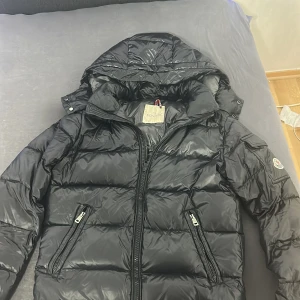 Moncler Maya  - Svart pufferjacka från Moncler med glansig finish, huva och två dragkedjeförsedda fickor. Jackan har Moncler-logga på ärmen och quiltad design. Perfekt för kalla dagar och har en riktigt clean look.
