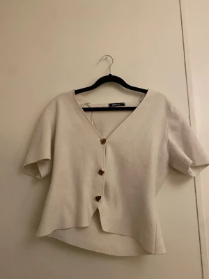 Vit kofta med v-ringning från Gina-tricot  - Superfin vit  kofta från Gina-tricot med v-ringning och korta ärmar. Koftan har snygga bruna knappar framtill och är tillverkad i ett mjukt, stickat material. Perfekt för lager-på-lager och enkel att styla till olika looks.