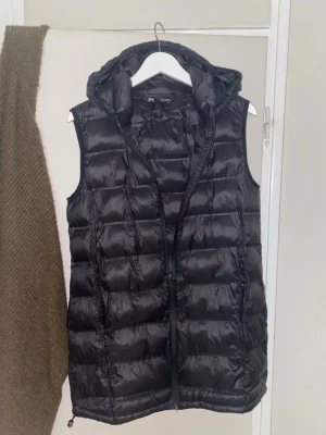 Svart dunväst från Zara med huva - Svart dunväst från Zara med quiltad design och praktisk huva. Västen har dragkedja framtill och två sidofickor med dragkedja. Materialet är glansigt och känns lätt men ändå värmande. Perfekt för lager-på-lager och chill stil.