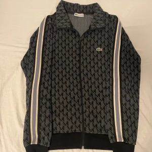 Svart mönstrad ziptröja Lacoste S - Snygg svart ziptröja från Lacoste i storlek S med geometriskt mönster och klassiska ränder längs ärmarna. Tröjan har dragkedja framtill, ståkrage och det ikoniska Lacoste-krokodilemblemet på bröstet. Perfekt för dig som gillar sportig och stilren vibe.
