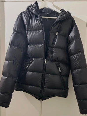 Svart Peak Performance pufferjacka S - Svart pufferjacka från Peak Performance med huva och flera dragkedjeförsedda fickor. Jackan har en stilren look med glansigt yttermaterial och breda, quiltade sektioner. Perfekt för kalla dagar och har en snygg, sportig vibe. Jackan har ett pyttelitet hål på baksidan, inget man märker! Köpt för 3000kr.