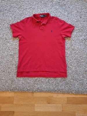 Polo Ralph Lauren röd pikétröja  - Riktig snygg röd pikétröja från Polo Ralph Lauren i storlek L. Custom fit. Använd fåtal gånger och skicket är bra. Den har ett hål på sidan(bild 7) som knappt syns och det påverkar inte tröjans skick i helhet. Den har en blå logga på vänstra bröstet. Skick: 7.5/10