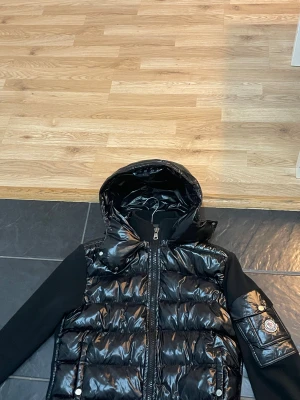  Moncler cardigan - Säljer en svart cardigan från Moncler med glansig finish och huva. Jackan har långa svarta ärmar i mjukt material, dragkedja framtill och en ikonisk Moncler-logga på ärmen. Fickor med tryckknappar och snygga quiltade detaljer ger jackan en modern vibe.