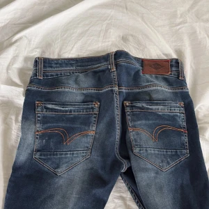 Lee Cooper Norris jeans med slitningar - Säljer ett par blå Lee Cooper Norris jeans med slim leg och snygga slitningar på benen. Klassisk femficksmodell med röda detaljer och kontrastsömmar. Perfekta för dig som gillar en smalare passform och lite tuffare look.