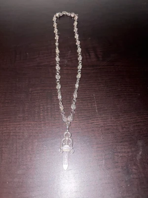 Chrome Hearts silver halsband med svärd - Unikt halsband från Chrome Hearts i silver med rundade länkar dekorerade med blommönster och ett detaljerat svärdhänge. Kedjan har en cool design och svärdet har ingraverad text. Perfekt för dig som gillar edgy och exklusiva smycken.