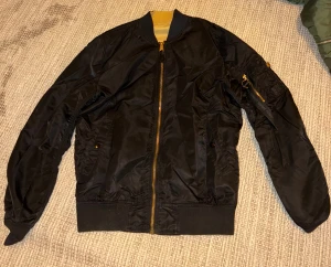 Svart och beige bomberjacka Alpha Industries - En svart och guldig bombar jacka. Man kan vända ut och in på den vilket gör den väldigt unik och cool. Från Alpha industries.