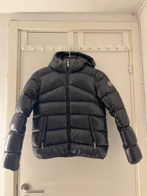 Svart pufferjacka från Colmar - Snygg svart pufferjacka från Colmar med glansig finish och huva. Jackan har två fickor med dragkedja, markerade sömmar och ett Colmar-märke på ärmen. Perfekt för kalla dagar och har en modern, sportig look.