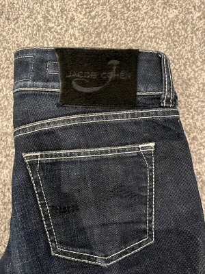 Jacob Cohen mörkblå jeans straight fit - Snygga mörkblå jeans från Jacob Cohen med klassisk straight fit och kontrasterande vita sömmar. Jeansen har fem fickor, exklusiv etikett av äkta hästpäls bak och detaljerad insida med mönstrat tyg. Perfekt för dig som gillar premiumkänsla och stilren design. Jeansen har även en egen parfymering. Dessa Jeans är för dig som vill ha en lyxig men även en stilren look! Jeansen är i mycket bra skick utan några skador/slitage! Nypris: 5500kr, mitt pris 1999kr. Pris kan diskuteras! 