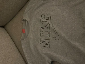 Grå Nike sweatshirt med tryck - Snygg grå sweatshirt från Nike med stort svart logotyp-tryck på bröstet. Tröjan har rund halsringning och är tillverkad i mjuk bomullsblandning. Perfekt för en sportig och avslappnad stil.