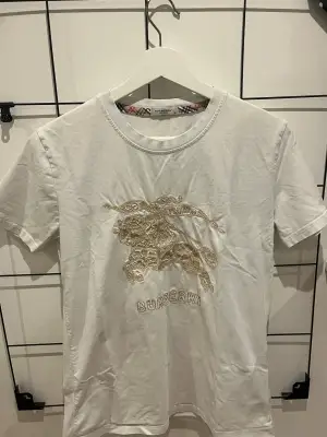 Vit t-shirt från Burberry med ikoniskt broderat ryttarmotiv och logga framtill i beige toner. Klassisk rund halsringning med rutigt band på insidan av kragen. Tillverkad i mjuk bomull och har korta ärmar med extra broderidetalj på ena ärmen.