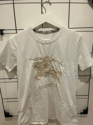 Vit Burberry t-shirt med broderi - Vit t-shirt från Burberry med ikoniskt broderat ryttarmotiv och logga framtill i beige toner. Klassisk rund halsringning med rutigt band på insidan av kragen. Tillverkad i mjuk bomull och har korta ärmar med extra broderidetalj på ena ärmen.