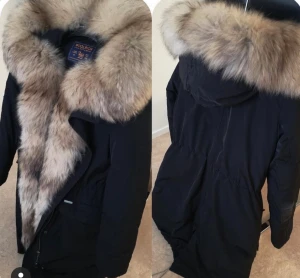 Svart parkajacka med päls från Woolrich - Säljer en svart parkajacka från Woolrich med stor fluffig päls runt huvan och kragen. Jackan har dragkedja framtill och flera fickor. Perfekt för kalla dagar med sin varma och stilrena look. Pälsen är ljusbrun och ger jackan en lyxig vibe.