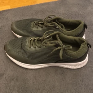 Olivgröna sneakers med vit sula - Säljer ett par olivgröna sneakers med vit och svart sula. Skorna har snörning och är tillverkade i mesh och syntetmaterial för en sportig look. Perfekta för dig som gillar stilrena och bekväma skor till vardags eller träning.