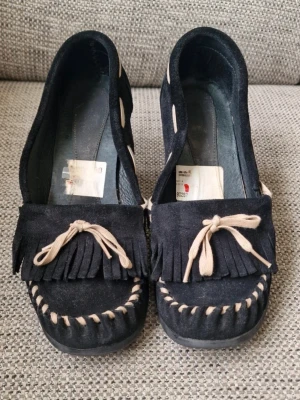 Skor med kilklack - Högklackade skor med inspiration från mockasiner. Använda men har fortfarande mycket kvar att ge. Finns lite skav bak på skorna (se bilder). Osäker på märke men på prislappen som delvis går att läsa så står det skohuset i alla fall. Stl 40