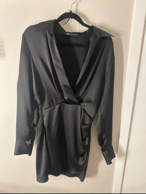 Svart klänning från Zara - Snygg svart kortklänning från Zara med omlott-design och djup v-ringning. Klänningen har långa ärmar med breda manschetter, krage och draperad midja som ger en cool siluett. Materialet är glansigt och känns lyxigt mot huden.