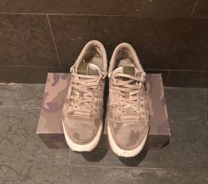 Valentino Garavani Rockrunner sneakers - Säljer ett par Valentino Garavani Rockrunner sneakers med grått och vitt kamouflagemönster. Skorna har snörning, platt sula och detaljer i mocka och skinn. Ikoniska nitar på hälen och meshpaneler ger en cool och sportig vibe. Kommer med originalkartong och dustbag. Pris går att diskutera!