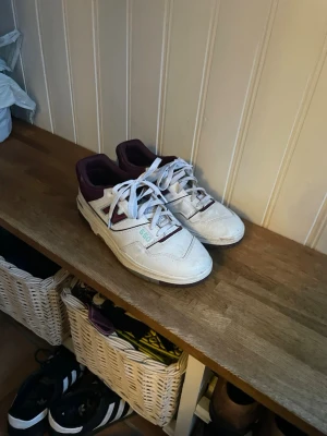 New Balance 550 vita sneakers - Säljer ett par New Balance 550 sneakers i vitt med mörkröda detaljer och snörning. Skorna har en klassisk låg siluett, rund tå och platt sula. Perfekt för dig som gillar retrostil och vill ha något som sticker ut lite extra. Väldigt bra skick, (lite smutsiga på bilden), storlek 45, pm för fler bilder, pris går att diskuteras 