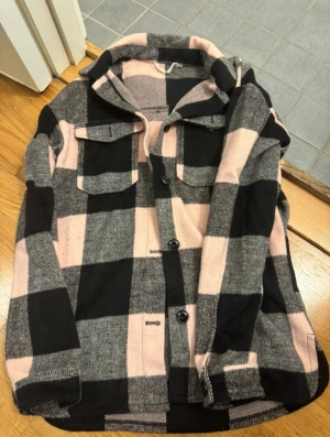Rutig overshirt i rosa och svart - Säljer en snygg overshirt med stora rutor i rosa, svart och grått. Jackan har krage, två bröstfickor med lock och knäppning framtill. Perfekt att slänga över en hoodie för en chill look. Passar dig som gillar streetstyle och lager på lager.