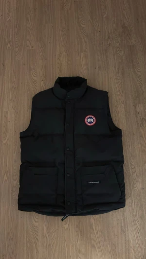 Svart dunväst från Canada Goose - Svart dunväst från Canada Goose med klassisk logga på bröstet. Västen har hög krage, två stora fickor framtill och knäppning med både dragkedja och tryckknappar. Materialet är polyester och dun, perfekt för kyliga dagar.