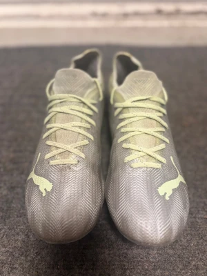 Puma Ultra grå fotbollsskor - Säljer ett par gråa Puma Ultra fotbollsskor med ljusgula detaljer och snörning. Skorna har en lätt ovandel i syntetmaterial och mönstrad textur för extra grepp. Klassisk rund tå och dobbar för spel på gräs. Perfekta för dig som vill ha stilrena och snabba skor på planen. Storlek 38.5. Ny pris 1500