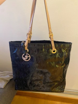 Michael Kors axelväska - Svart axelväska från Michael Kors med glansig finish och MK-logga i mönstret. Väskan har ljusbeige justerbara axelremmar i skinn och guldfärgade detaljer. Insidan är fodrad med logotypmönster och har flera fack för småsaker. Perfekt storlek för allt du behöver🌸