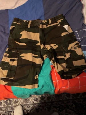 Camo cargoshorts med fickor - Säljer ett par cargoshorts i klassiskt camouflage-mönster med flera stora fickor på sidorna. Shortsen är i grönt, beige och svart och har bälteshällor samt knappstängning. Perfekta för dig som gillar streetwear och vill ha en avslappnad stil.