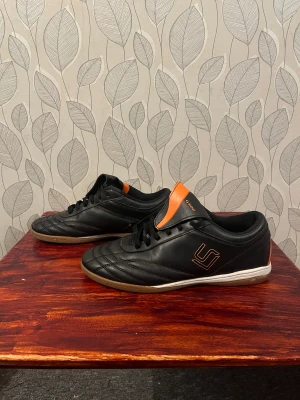 Svarta fotbollsskor från Soc - Säljer ett par svarta fotbollsskor från Soc med orangea detaljer och vit sula. Skorna har snörning och är tillverkade i syntetmaterial med klassisk låg profil och rund tå. Perfekta för inomhusspel och har en stilren design med logga på sidan och hälen.