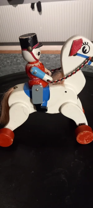 Trädhäst 1950  - Västen är röd med svarta knappar framtill och har en klassisk, rundad form. Den är tillverkad i trä och har målade detaljer. Västen bärs av en leksaksfigur med blå ärmar och svart hatt, vilket ger en cool och retro look.