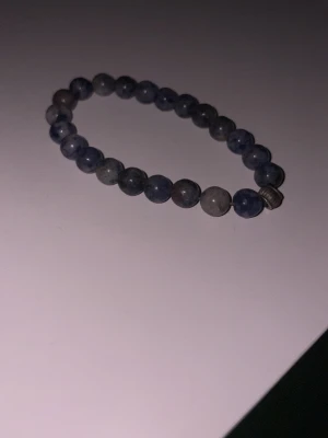 Två pärlarmband med stenar - Säljer två handgjorda pärlarmband. Ett med blå och grå runda stenar och metallpärla, och ett med beige, orange och gulmelerade stenar samt en grå kubformad pärla. Perfekt för dig som gillar unika accessoarer och vill ha något som sticker ut.