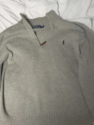 Grå stickad tröja Polo Ralph Lauren - Snygg grå stickad tröja från Polo Ralph Lauren med halv dragkedja och broderad blå logga på bröstet. Tröjan har ribbade muddar och hög krage, perfekt för kyliga dagar. Klassisk och stilren design som funkar till det mesta. Är en Xl i storlek men sitter mycket mer som en L