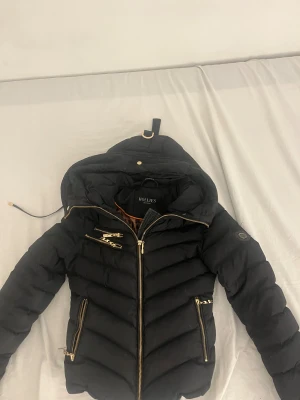 Svart pufferjacka Hollies med leopardfoder - Snygg svart pufferjacka från Hollies med guldiga dragkedjor och detaljer. Jackan har huva och ett coolt leopardmönstrat foder på insidan. Saknar dock luvan