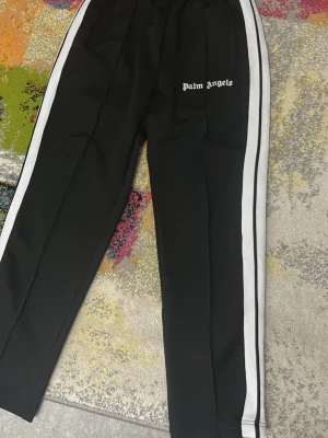 Palm Angels svarta mjukisbyxor M - Svarta mjukisbyxor från Palm Angels med vita sidoränder och logga framtill. Byxorna har elastisk midja, raka ben och dragkedja vid benslut. Snygg för att hänga med personer och bra för alla väder