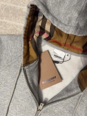 Burberry zip hoodie grå – dam M / herr S - Grå Burberry zip hoodie med klassiskt rutigt foder i huvan. Broderad Burberry-logga framtill. Brand New – aldrig använd, i perfekt skick utan fläckar eller hål. Mjuk och bekväm kvalitet. Storlek M i dam och S i herr (unisex).