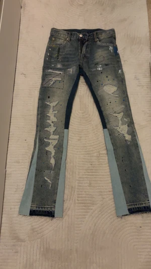 Unika blå jeans med patch och färgstänk - Säljer ett par riktigt coola jeans från MSL med patchade detaljer, färgstänk och slitningar framtill. Byxorna har ljusblå paneler längs benen och fransade benslut. De har en rak passform och klassisk femficksdesign. Perfekt för dig som gillar streetstyle och vill sticka ut.