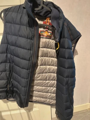 Parajumper väst - Säljer en blå dunväst från Parajumpers i modellen Super Lightweight. Västen har quiltad design, dragkedja och detaljer i grått på insidan. Perfekt för lager-på-lager och har en sportig look. Köpt för 4500kr. Säljes för 1600kr. Storlek M men passar som S