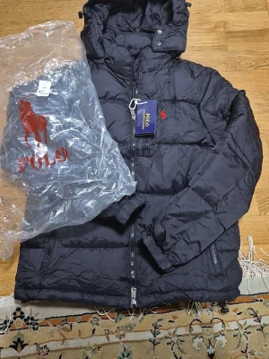 Svart pufferjacka Polo Ralph Lauren - Svart pufferjacka från Polo Ralph Lauren med röd broderad logga på bröstet. Jackan har huva, två sidofickor med dragkedja och quiltad design. Materialet är syntet och polyester, perfekt för kalla dagar. Stängs med dragkedja framtill och har elastiska muddar vid ärmslut.