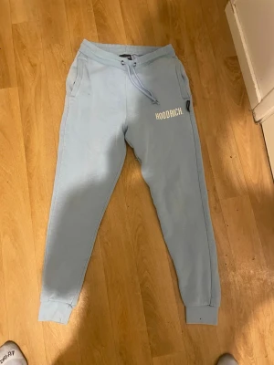 Ljusblå mjukisbyxor från Hoodrich - Säljer ett par ljusblå mjukisbyxor från Hoodrich med snörning i midjan och ribbade muddar vid bensluten. Byxorna har två sidofickor och Hoodrich-logga tryckt på ena benet. Perfekta för chill eller träning, riktigt sköna och stilrena.