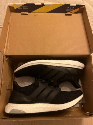 Adidas Ultraboost svarta sneakers - Svarta Adidas Ultraboost sneakers med stickad ovandel och klassiska tre ränder på sidan. Skorna har en vit Boost-mellansula för extra komfort och en svart yttersula med greppvänligt mönster. Snörning framtill och platt sula. Perfekta för dig som gillar stilren och sportig look.