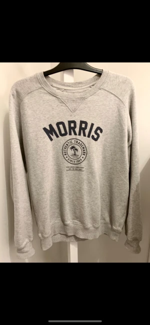 Grå sweatshirt från Morris - Säljer en grå sweatshirt från Morris med tryck i mörkblått framtill. Tröjan har rund halsringning, ribbade muddar vid ärmslut och nederkant samt lång ärm. Perfekt för en avslappnad och stilren look.